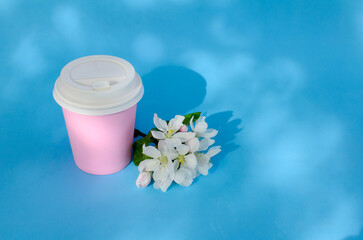 Obraz na płótnie Canvas Pink paper cup for coffee and other beverages closed with white lid, spring bouquet of apple blossoms with shadows on blue background. top view. Natural eco packaging.