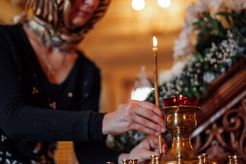 Cropped photo of middle-aged woman wearing headscarf, putting burning candle on golden stand with candlesticks in orthodox church. Faith in God, religion, ceremony, praying, holiday, commemoration.