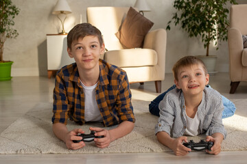 Two joyful boys lie on the floor in a room playing video games with joysticks