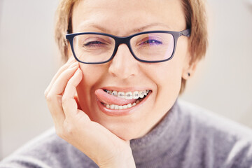 Stylish and beautiful young blonde woman in glasses and a gray sweater in braces and smiling on a white background, dentist orthodontist concept