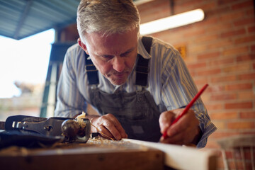 Mature Male Carpenter In Garage Workshop Marking Wood With Pencil