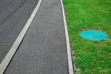 Asphalt road with tarmac footpath with stone curb and septic tank manhole on green lawn, nobody.