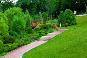iron ground lantern and poli garden lighting of park with stone stairs and path paved tiles with drain lattice in park among plants, evergreen bushes and trees on slope hill green lawn, nobody.