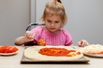 Little girl making pizza. Small child cooking meal, sitting at the kitchen table at home. Kid preparing food.