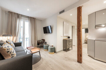 Living room with three-seater sofa, wooden coffee table, balcony with curtains and net curtains, wooden column and kitchen with gray furniture