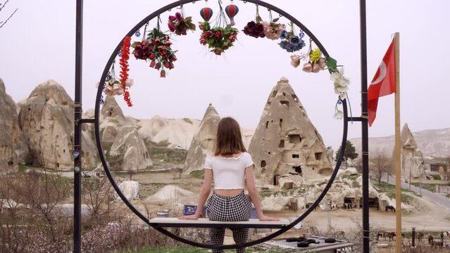 Lonely Young Caucasian Woman Sits On Fower Decorated Bench and Looking to StonE Valley in Cappadocia Turkey Flag. Girl at Vacation Travel to Rocky Mountain. Rear Back View. Medium Shot