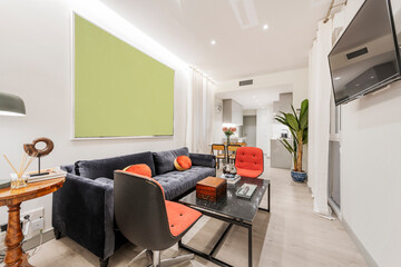 Living room with three-seater blue sofa, red cushions, black marble coffee table and red armchairs on wooden floor, flat screen TV on the wall and decorative plants