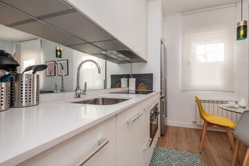 apartment kitchen with white lacquered cabinets, white stone countertop with built-in sink, mirror on the wall and dining table in front