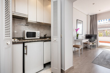 Small kitchen of an apartment with a small fridge under the gray granite countertop and a microwave above it, gray wooden doors and access to a living room with access to a nice urban terrace