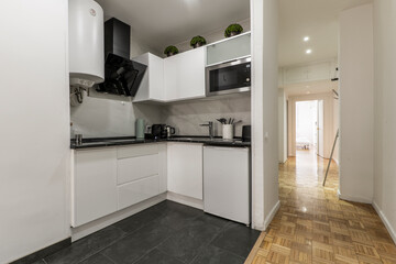 Fototapeta na wymiar Corner kitchen with white wooden furniture, integrated appliances, black countertop and combined floor of black stoneware and oak parquet floors