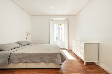 Fototapeta na wymiar Bedroom with double bed with gray bedspread, white wooden chest of drawers and balcony with wooden shutters