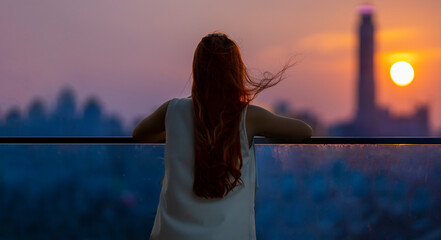 Woman looking and enjoying the sunset view from balcony with the sun setting behind skyscraper in busy urban downtown with loneliness for solitude, loneliness and dreaming of freedom lifestyle