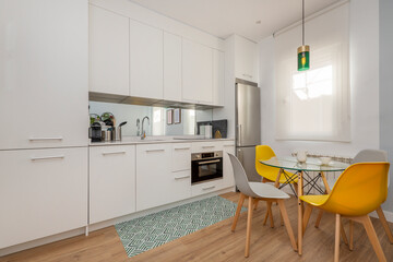 Kitchen open to living room with white cabinets and white stone countertops, matching appliances and glass dining table with gray and yellow resin and wood chairs