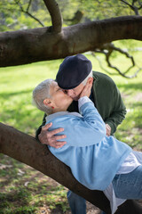 Romantic elderly husband and wife kissing in park. Happy man and woman hugging kissing when woman lying on thick tree branch and enjoying rest. Love, relations and active rest of aged people concept