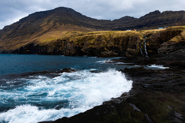 Nordic coastline with breaking ice-blue ocean on black rocky beach, cliff with waterfall and mountain riff in background, cloudy grey sky