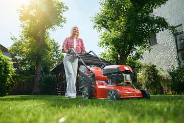 Low angle view of beautiful caucasian woman with blond hair using electric lawn mower for cutting grass on back yard. Pretty female in casual wear using modern equipment for work at garden.