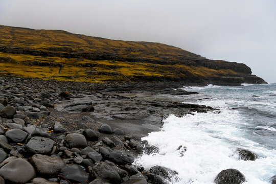 Rocky nordic black beach with cold stormy breakers, grass covered cliff in background, misty cloud cover