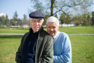 Portrait of senior couple on grassy field. Smiling man in cap and woman resting in park, woman leaning mans shoulder hugging him, both looking at camera. Relations, active rest of aged people concept