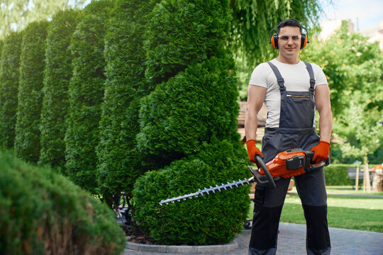 Competent caucasian gardener smiling and looking at camera while standing on back yard and holding hand electric trimmer. Man using gardening tool for cutting hedge. 