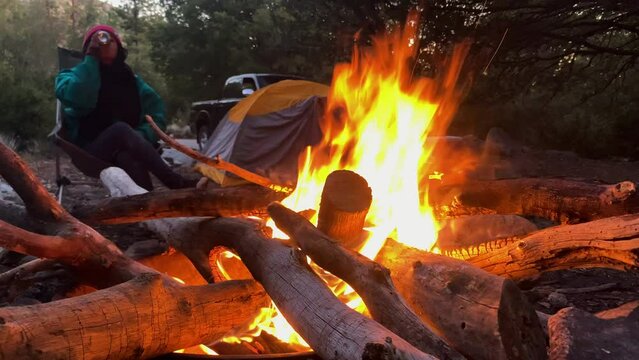 young trekker female woman resting in front of bonfire while drinking can of beer, camping tent equipment for long trail hike