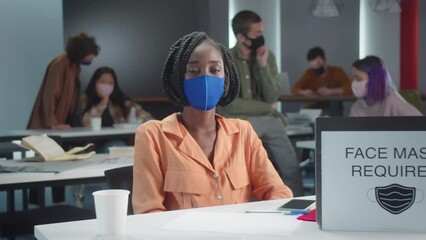 A young african professor is sitting in a blue protective mask with her students in the classroom and looking at the camera