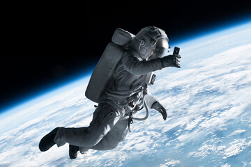 Female astronaut having a video call on her phone while performing spacewalk in open space, Earth in the background - obrazy, fototapety, plakaty
