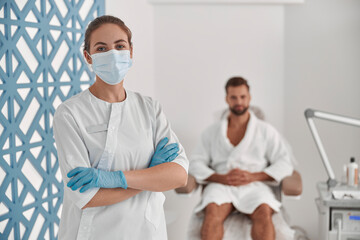 Portrait of professional beautician in protective mask and gloves near patient in cosmetologycal clinic