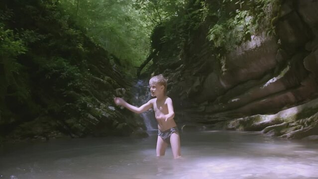 A child splashes in a pond near a waterfall. CREATIVE. A little white boy is sitting in the lake. White-haired boy playing in the pond