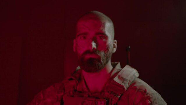 Serious mn in camouflage uniform standing in dark room. Front view of man with scratches on his face holding shotgun, posing for camera after playing airsoft. Game, emotion concept