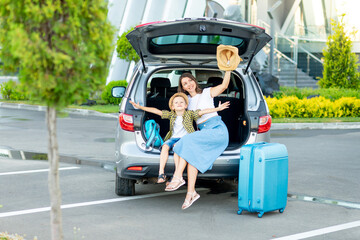 a mother with a child son and a blue suitcase are going on vacation or a trip sitting in a car in front of the airport and waving a hat