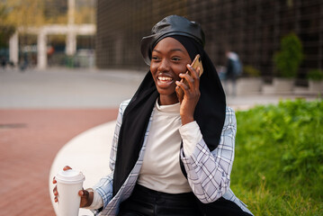 Cheerful black woman with takeaway coffee speaking on smartphone