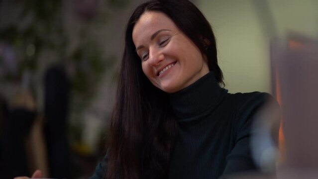 Young beautiful Caucasian thoughtful brunette girl in dark jumper cute smiling, sit in cafe. Close-up portrait of happy woman, indoors view. Slow motion.