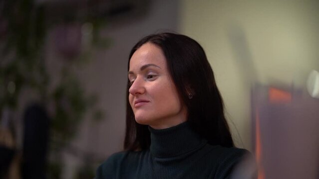 A thoughtful young brunette girl in dark jumper is sitting in a coffee shop, looking around, waiting and sad on blurred background. Close up portrait of alone woman slow motion.
