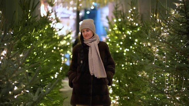 One European woman with a long plait of black hair in winter hat, a fur coat walks along the alley between Christmas trees decorated with glowing garlands on street. Holiday concept.