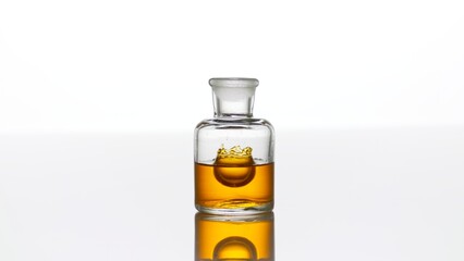 Drop of argan oil falls down into clear old fashioned bottle with yellow transparent liquid on white background making crown inside it | Abstract cosmetics formulation concept