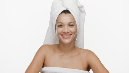 Young cheerful African American woman wrapped in towel and with hair drying towel on her head smiling wide for the camera on white background