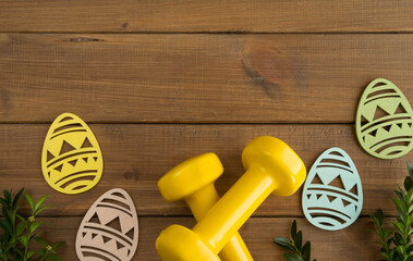 Two heavy dumbbells, boxwood branches and decorative Easter eggs. Healthy fitness lifestyle composition, gym workout and training concept. Fit flat lay with copy space on wooden background.
