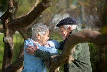 Side view of aged couple resting under trees. Smiling woman looking at man with joy and man hugging her, both standing leaning thick tree branch in park. Love, relations of aged people concept