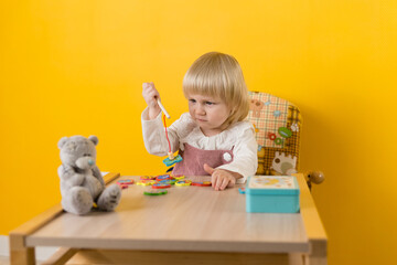 A girl in dress is sitting at table against a yellow wall and enthusiastically playing fishing with a magnetic toy. Development of motor skills, attention, coordination, perception of shapes, colors