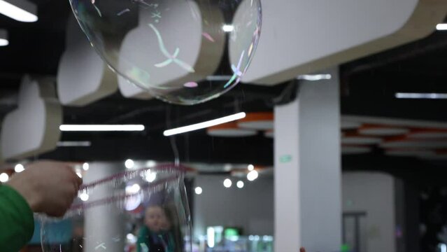 In A Closed Amusement Park, A Presenter In A Green Suit Inflates Huge Soap Bubbles For Children. Little Baby Pens Happily Reach Out To Burst Giant Soap Bubbles, Sweat And Many Small Ones