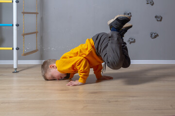 A Caucasian boy in an orange hoodie is standing on his hands with his legs raised up, frozen, the child is dancing a break dance to the room