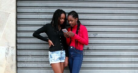 Candid black teens girls using cellphone, adolescent girl in shock gossiping with friend