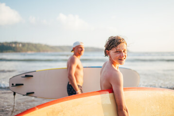 Smiling teen boy with surfboard go to the sea for surfing. He have a winter vacation and enjoying a beautiful sunset light with father on Sri Lanka island. Family active vacation concept.