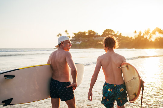 Father with teenager son walking with surfboards by sandy ocean beach with palm trees on background lightened with sunset sun They have conversation and enjoy summertime Family active vacation concept