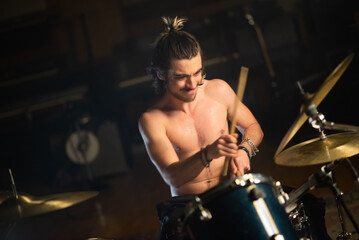 Fototapeta na wymiar Enthusiastic male musician playing drum set in garage. Young Caucasian man sitting shirtless and training or recording drums in studio. Drummer training concept