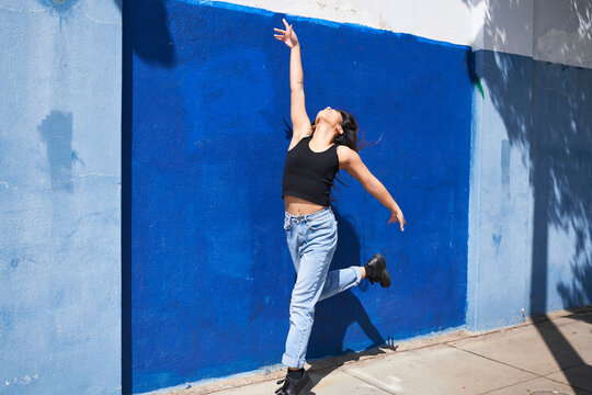 Woman dancing and leaping in front of blue wall outdoors arm up