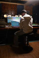 Rear view of young sound engineer looking at laptop in sound recording studio. Caucasian man sitting at mixing console panel. Creating music concept