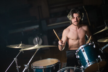 Fototapeta na wymiar Excited young rock drummer playing drum set in studio. Man with moustache sitting shirtless at drum set and rehearsing or recording music. Drummer training concept