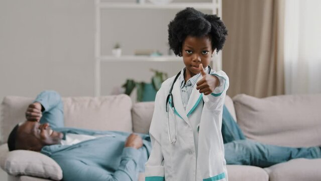 Little african american daughter playing with dad father patient lying on couch kid girl pretending to be doctor looking at camera smiling showing thumb up no problem excellent health approval gesture