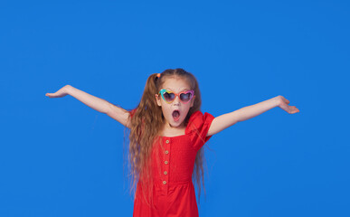 Portrait of surprised cute little toddler girl in the heart shape sunglasses. Child with open mouth having fun isolated over blue background. Looking at camera. Wow funny face